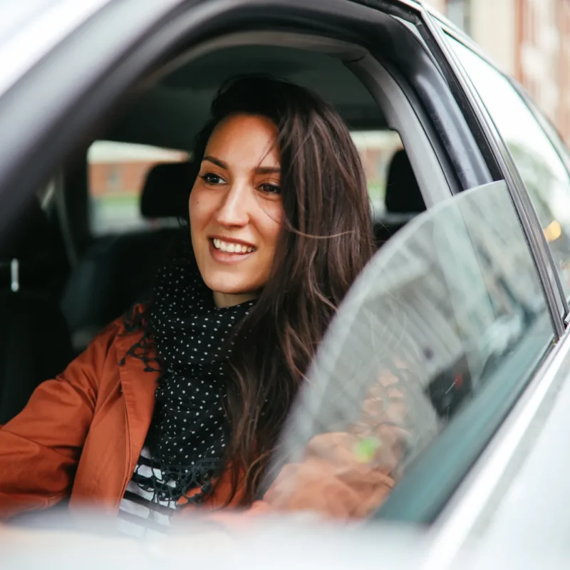 A smiling woman in an orange jacket sits behind the wheel of a silver car with the driver's window rolled down on a city street.