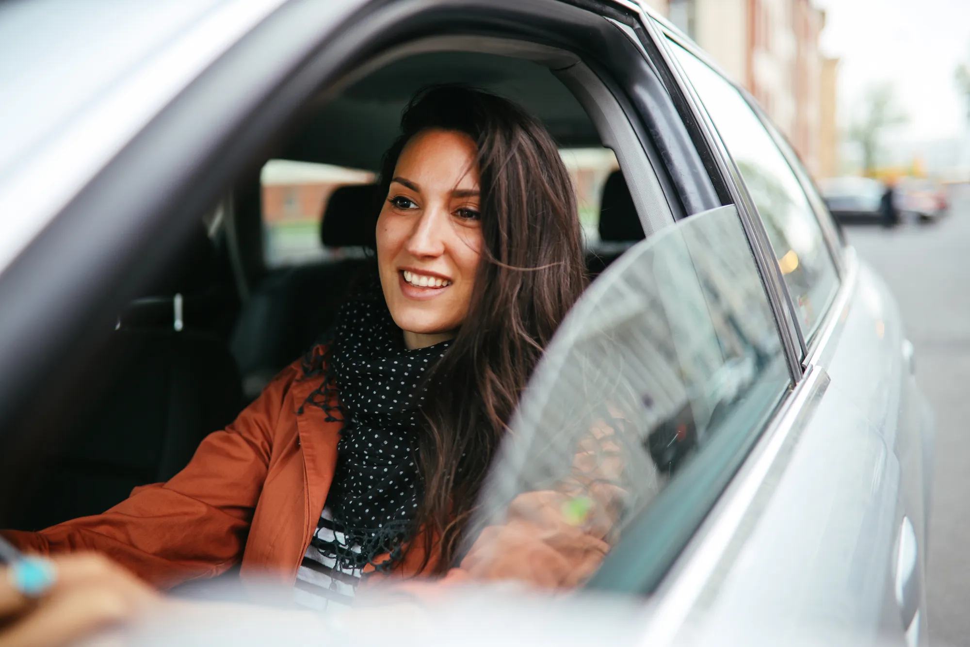 A smiling woman in an orange jacket sits behind the wheel of a silver car with the driver's window rolled down on a city street.
