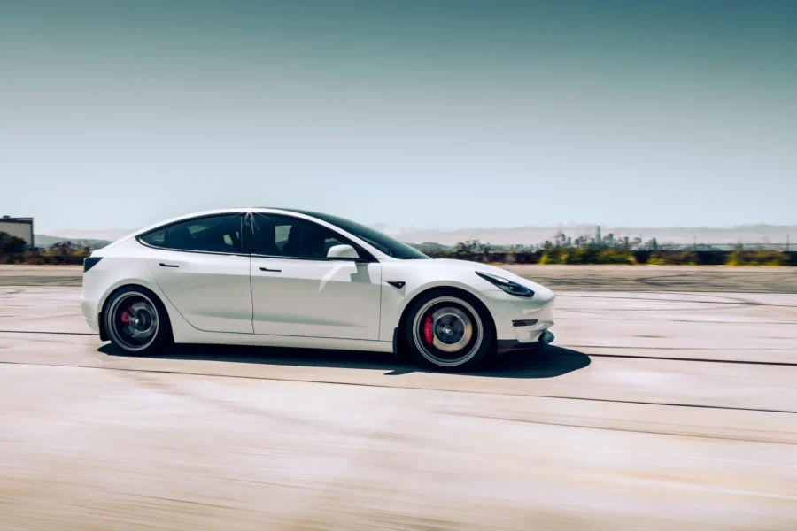 A white Tesla Model 3 sedan with dark alloy wheels and red brake calipers drives at speed across open pavement with a city skyline in the distance.