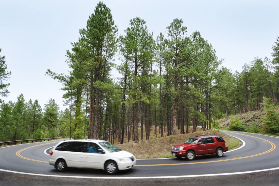 A white minivan and a red SUV pass each other on a hairpin curve of a pine-lined mountain road.